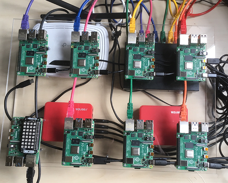 A picture of a cluster of eight Raspberry Pis in two rows of four which are mounted on a Perspex sheet. The one in the bottom left-hand corner has a matrix of LED lights on top of it. Each Raspberry Pi has a network cable and USB cable, for power, connected to it. Two USB hubs, a network switch and router are also shown under the Perspex sheet.